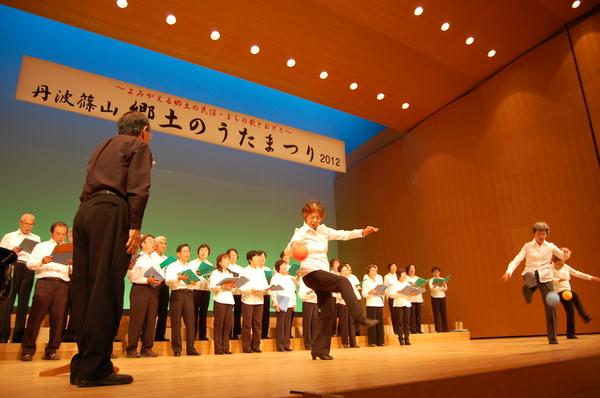 舞台上で、合唱団の歌に合わせて、3名の女性が「まりつき」をしている様子の写真