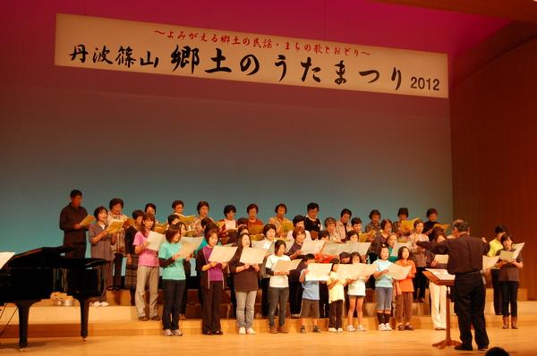 小学生ぐらいの子どもから大人までが一緒に舞台の上に並んでおり、歌詞を見ながら指揮者に合わせて、合唱している様子の写真