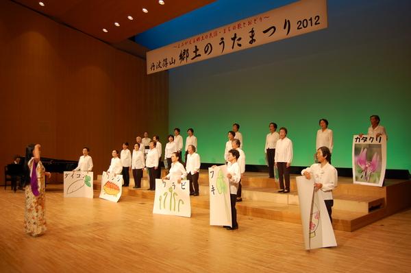 ダイコン、イノシシ、ワラビ、フキなどのイラストを大きな紙に描いたものを見せながら、歌を歌っている様子の写真