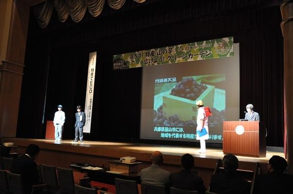 壇上のスクリーンの前で、赤いランドセルと黄色い帽子を被った小学生の恰好をした大人が篠山特産の「丹波黒大豆」について説明しており、左端の方に立っている男性2名、右端に司会者1名が説明を聞いている写真