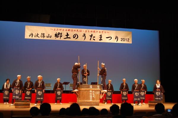 舞台の中央に大きな酒樽が置かれており、「丹杜」の文字の入った前掛けをかけた酒蔵の作業をする衣装を着けた男性が酒樽に設置された台に竹をもって立ち、舞台上に立ってい同じ衣装の男性と一緒に声を合わせて歌を歌っている様子の写真