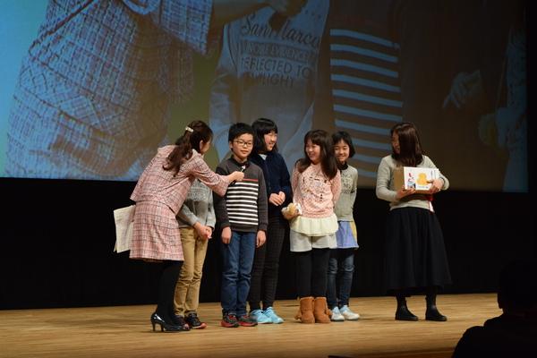 舞台のスクリーンの前でインタビューを受けている眼鏡をかけた男の子と同じグループの女の子らの写真