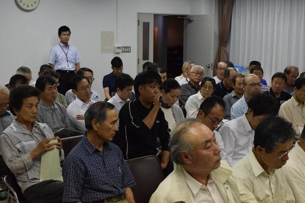 参加者が座っていて一人の男性がマイクを持って話をしている写真