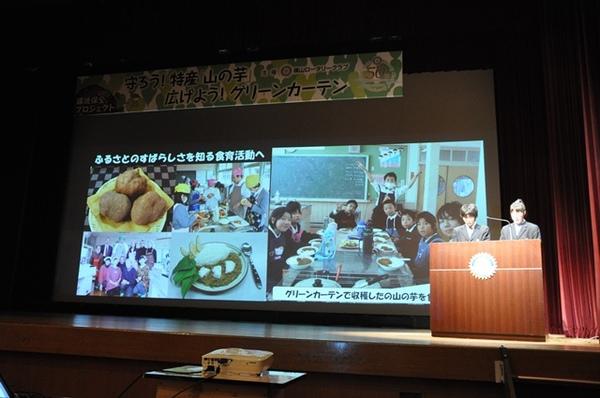 壇上のスクリーンに子どもたちが食育活動をしている様子を2名で説明している写真