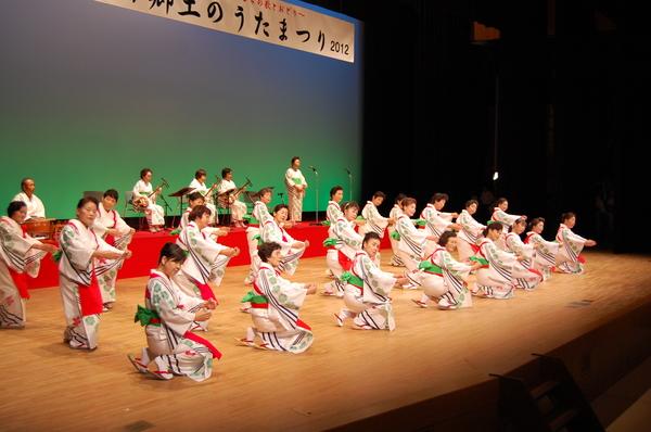白地に緑の花模様が入った銀杏に赤い前掛けを付け着物を着た踊り子が、両手を合わせて丸い形を作って右に顔を傾けるポースを取って踊っている様子の写真