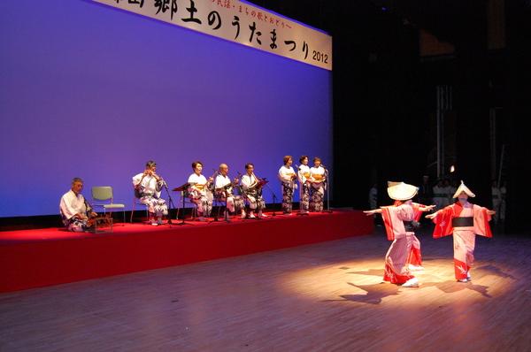 三味線や笛の演奏に合わせ3名が歌を歌っており、舞台の中央では、編み笠を被り、白地に赤い袖の着物を着た踊り子3名が踊っている様子の写真