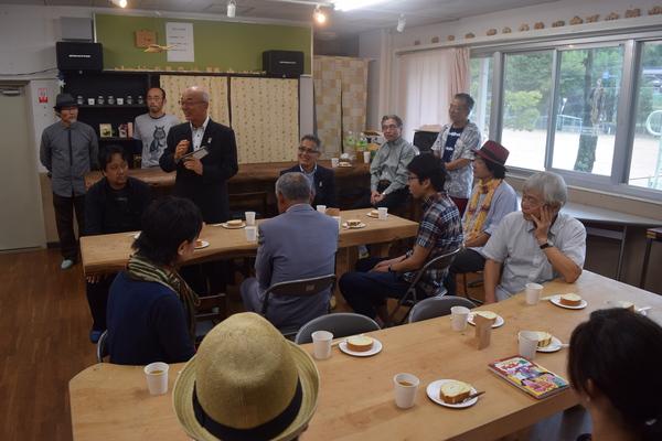 木で作られたテーブルにケーキとお茶が置かれてあり、その周りに10名ほどの人が座り市長が立って話をしている写真