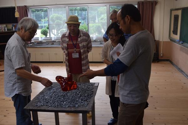 中央のテーブルに赤色のオブジェが置かれていてそれを5名の男女が見ている写真