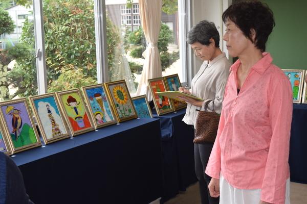 額縁に入った絵が窓際に8点飾られていて、それを2人の女性がメモをしながら見ている写真