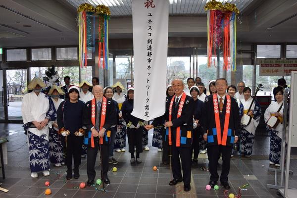 「祝 ユネスコ創造都市ネットワーク認定」の垂れ幕と2つのくす玉が割れており、法被を着た市長と白髪にあごひげの男性と関係者とその後ろにデカンショの踊り子や演奏者が一緒に写っている写真