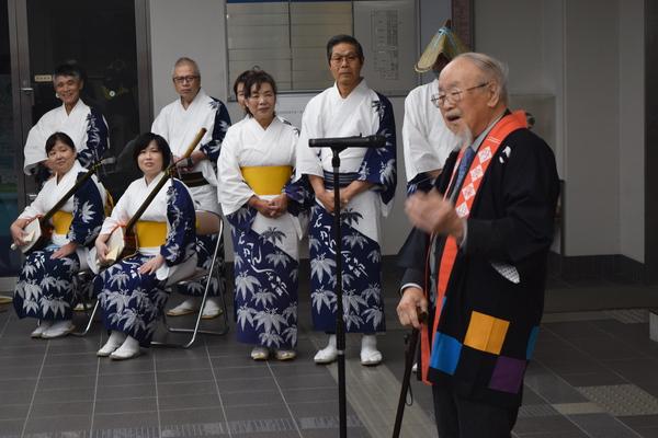 白髪に、あごひげのある男性が、スタンドマイクの前で話をしており、デカンショ踊りの演奏者や踊り子の皆さんが真剣な表情で話を聞いている様子の写真