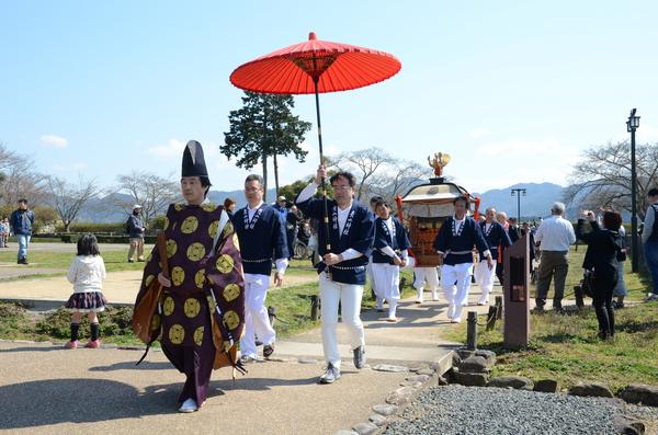 紺に法被を着た男性が赤い舞踏傘を手に持ちその後ろでは神輿を運んでいる写真