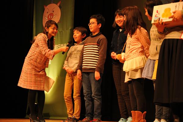 舞台の上でインタビューを受けているパーカーを着た男の子と同じグループの女の子らが笑顔の写真