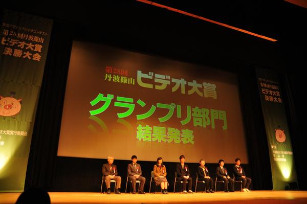 ビデオ大賞グランプリ部門の結果発表をスクリーンの前に座って発表を待っている7名の写真