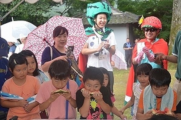 男の子3人がスイカの早食い競争に参加しスイカにかぶりついており、うしろには誰が早いが覗き込んでいる子やスイカのかぶりものをしている男性が写っている写真