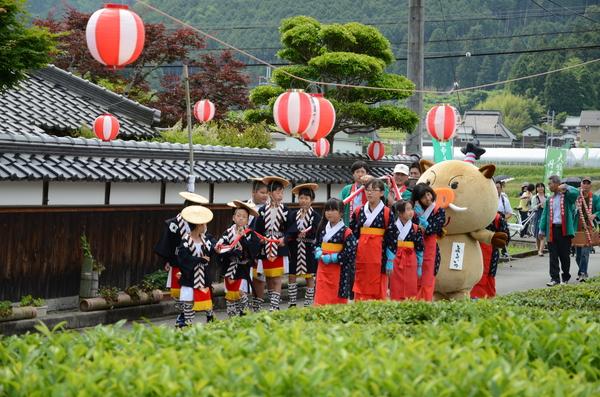 茶畑の横の道路を茶娘の衣装を着た女の子とその後ろに「まるいの」が歩いており、その隣に頭に傘を被っている男の子の行列が歩いている様子が写っている写真