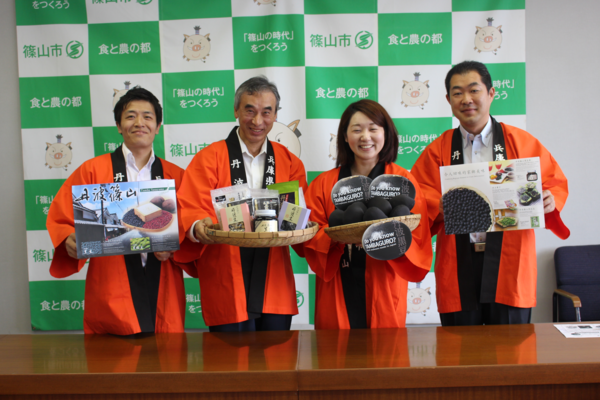 オレンジ色の法被を着て黒豆についての説明のパネルや黒豆の加工食品を持っている4人の職員の写真