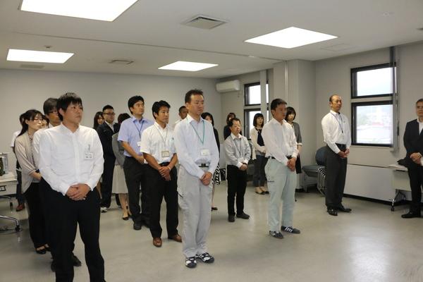 職員たちが並んで立ち手を前に組み話を聞いている写真