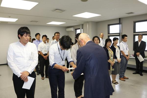 男性職員が市長から辞令交付を渡されて頭を下げ受け取っているのを周りの人達がみている写真
