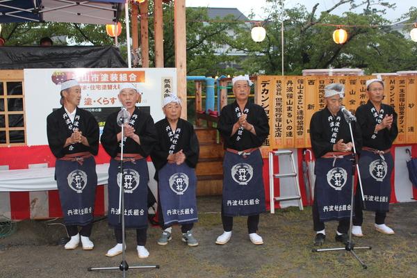 黒い法被に手ぬぐいを頭に巻き、エプロンをつけた男性6名が、手をたたきながら歌っている写真