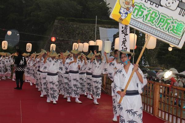市役所のプラカードを持った男性を先頭に、浴衣と編み笠を被った男女が躍っている写真