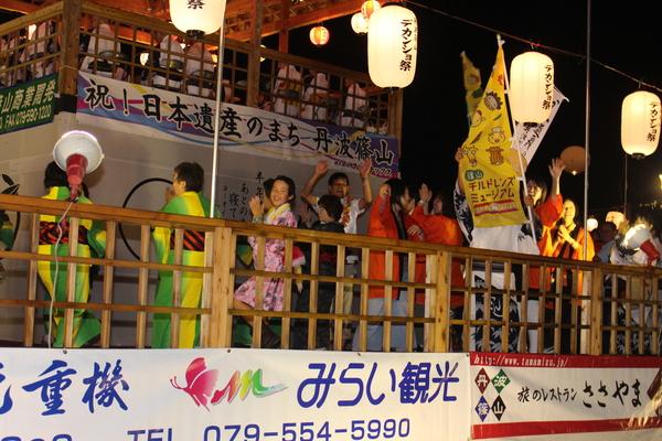 やぐらの上で笑顔で踊る、浴衣や、衣装を着た老若男女の写真