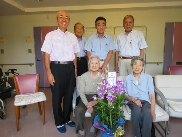 お祝いの花が置いてあり、おばあちゃんが2人座っていて、市長と関係者が後ろに立ち記念撮影している写真