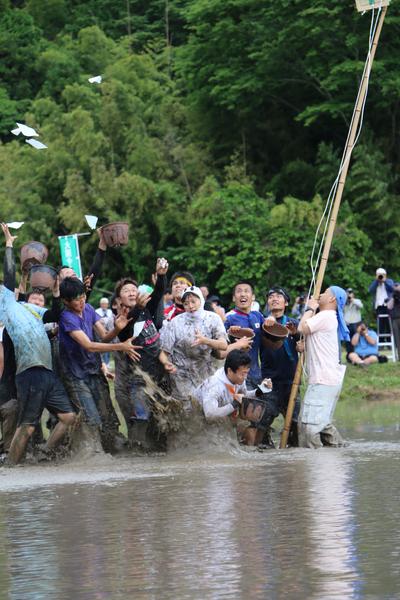 水の入った田んぼの中で、男性達が鉢のようなものを持っており、どろんこになりながら、上から飛んできた白い紙をキャッチしようとしている様子の写真