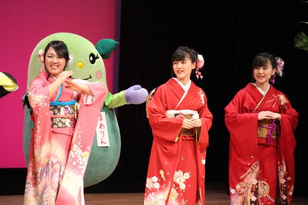 舞台の上で、振袖を着た女性3人が胸の前に手を置いて恋ダンスを踊っており、「まめりん」が後ろに写っている写真