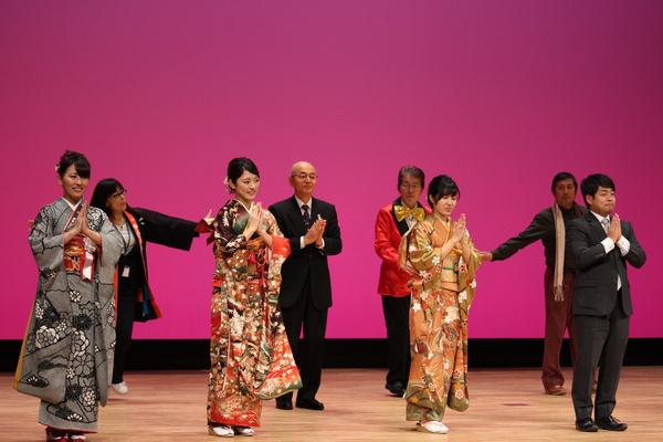 振袖を着た女性3人とスーツの男性が前方にてを両手を合わせ立ち、その後ろに市長も同じポースで立っており、赤いタキシードの男性、法被を着た女性も写っている写真