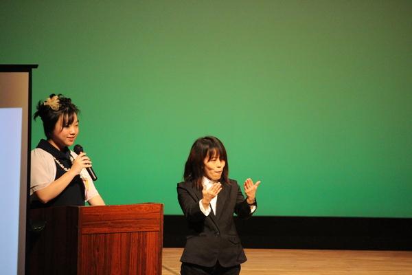 演説台の前にマイクを持った女性が話をしており、その横に手話通訳者の女性が同時通訳をしている様子の写真