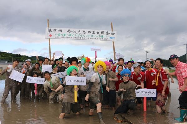 「第9回 丹波ささやま大田動会」と書かれた横断幕の前で、大会の参加者がどろんこになってチーム名の書かれたプレートを持って写っている集合写真