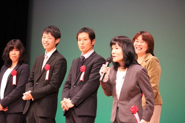 胸に胸章を付け、スーツを着た男性と女性が数名舞台上に立っており、右側の女性がマイクを持って話をしており、となりにいる女性が話を聞いて笑っている様子の写真