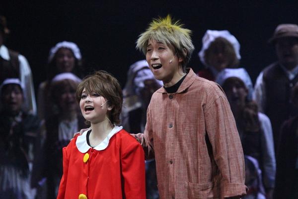 赤の衣装の女性と茶色に金髪の男性が歌っていて、後ろに白い衣装を着た方が並んで歌っている写真