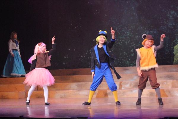 ピンクの衣装、青い衣装、黄色と茶色の衣装を着た3人が左手の指を上に指している写真