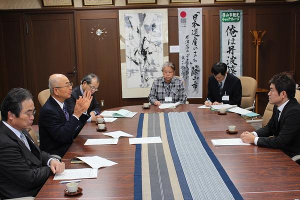 会議テーブルを囲み着席する市長と、菟原 元彦委員長や関係者の人達が資料に目を通したり話を聞いたりしている写真