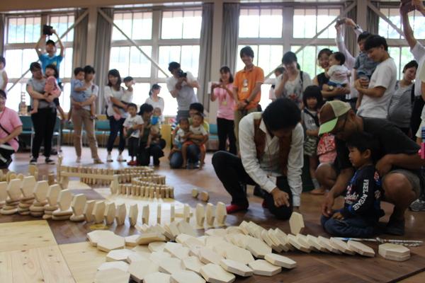 木育キャラバンで講師が男の子の前で六角形の木材を立てて指さしているのを見ている親子連れの写真