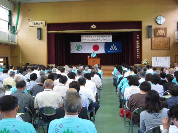 畑小学校創立139周年記念式典が行われている体育館の壇上で市長が挨拶をしているのを座って聞いている人々の写真