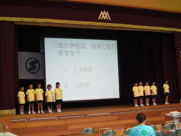 畑小学校創立139周年記念式典が行われている体育館の舞台でスクリーンの両脇に生徒が5名ずつ並んでいる写真