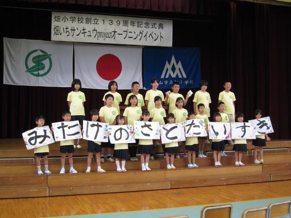 畑小学校創立139周年記念式典が行われている体育館の舞台に生徒たちが3列に並んでおり、前列の生徒たちが一文字ずつ書かれている画用紙を持って写っている写真