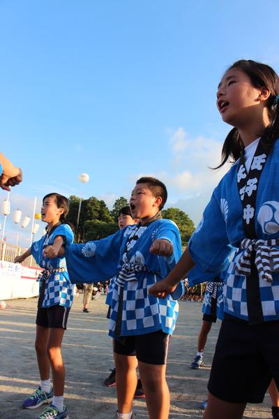 氏子中とかかれた水色のハッピをきた女の子の間に男の子が入り3人が横列になり手をグーにして両手をのばし声をだしている写真