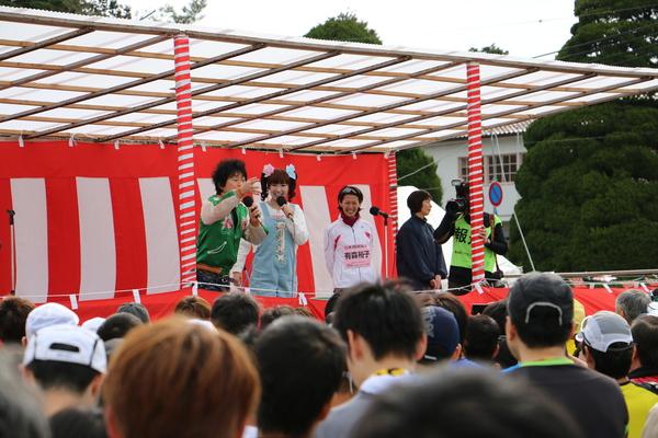 紅白幕の中で芸人のかつみ・さゆりさんがマイクを持って参加者に話をしている写真