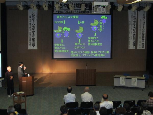 胃がんリスク検診の文字や胃の絵が描かれたスクリーンを見ながらスーツを来た男性が説明している写真