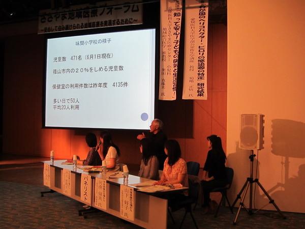 味間小学校の様子 保健室の利用件数は昨年度4135件と書かれているスクリーンを見ながらマイクを持った女性が説明している写真