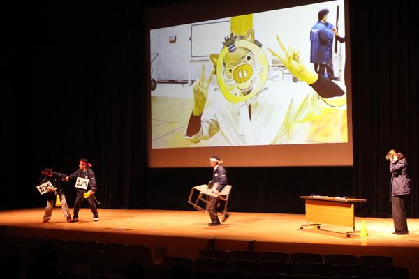 舞台で職員の方が劇をしていてスクリーンでは人の顔に動物の絵が写っている写真