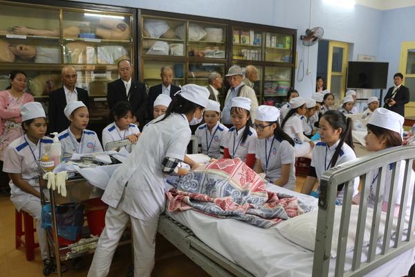 手前に立っている女性がベッドの上に置かれたシーツを触っていて、ベッドの周りに9名の学生たちが座っている写真