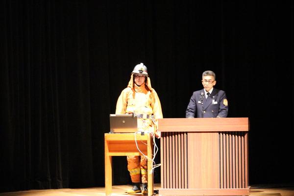消防服を着ている男性はパソコンの前に、制服を着た男性は壇上でお話している写真