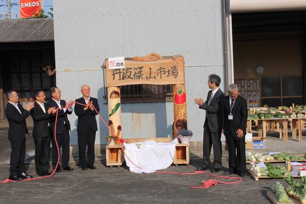 柱が二本あってその上に丹波篠山市場と書かれている木の作り物があり、その横で男性6名が拍手をしている写真