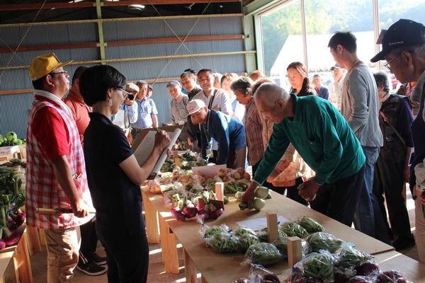 机の上に玉ねぎやいんげんなどの野菜が置かれていて、来場者達が野菜を手に取ったりしながら見ている写真