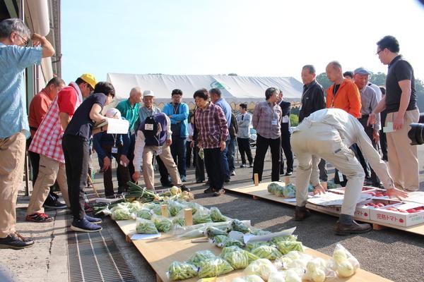手前には玉ねぎ、トマトなどの野菜が置かれその後ろに20名ほどの来場者達が立っている写真